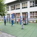 Grupa dzieci bawi sie na placu zabaw. W głębi budynk szkolny koloru biało-szarego