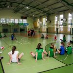 Grupa dziewcząt w sporowych strojach siedzi na podłodze koloru zielonego w hali sportowej