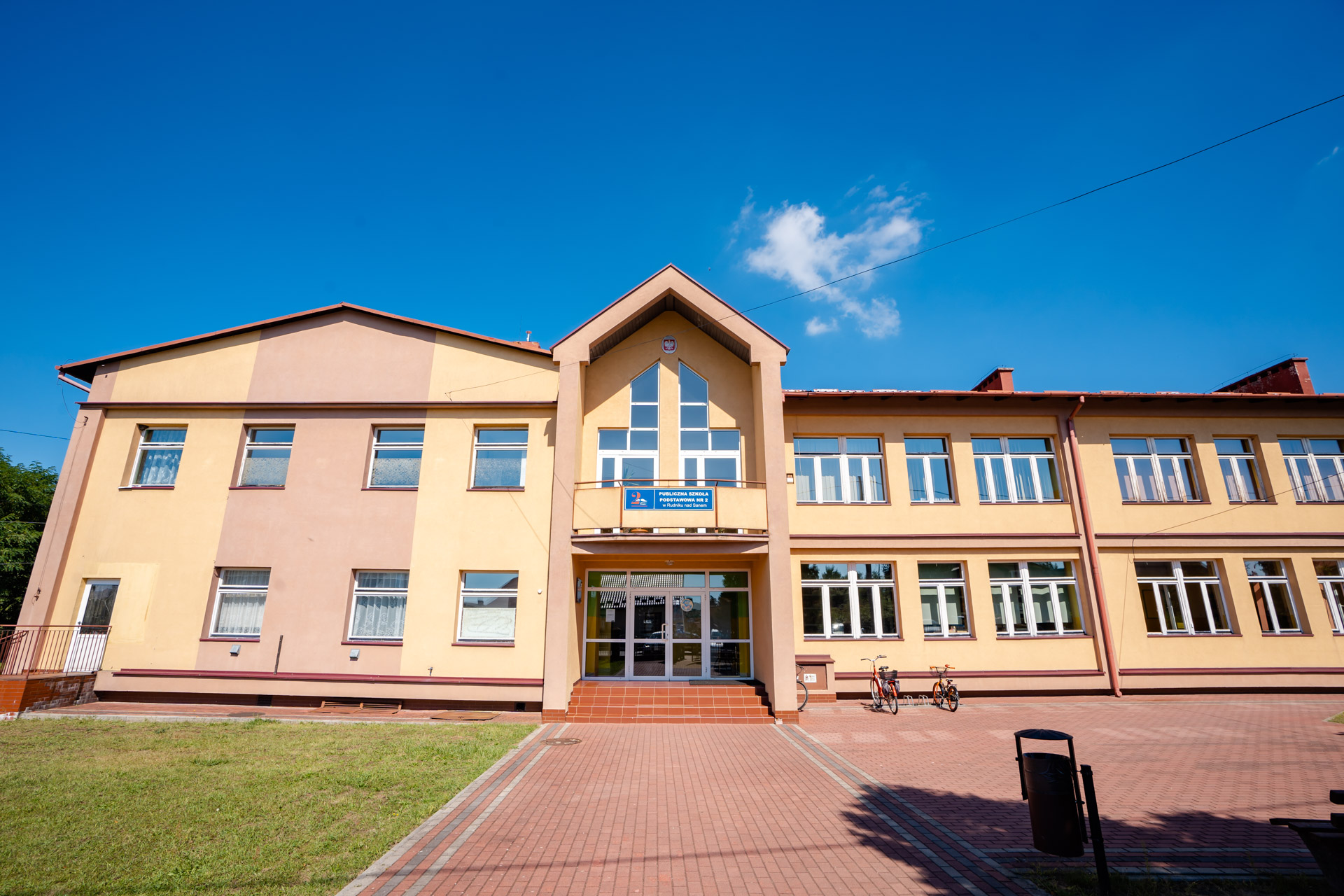 Jednopiętrowy budynek szkoły, na środku przeszklone wejście. Elewacja koloru różowego i żółtego
