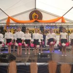 Grupa dziewcząt w białych bluzkach i czarnych spódniczkach z biało-różowymi ozdobami tańczy na scenie.