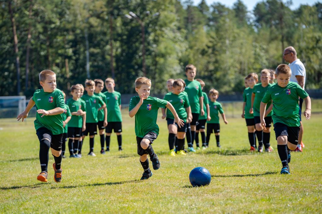 Chłopcy w zielono-czarnych strojach trenują na trawiastym boisku. Z boku stoi mężczyna w granatowych spodeńkach i białej koszulce