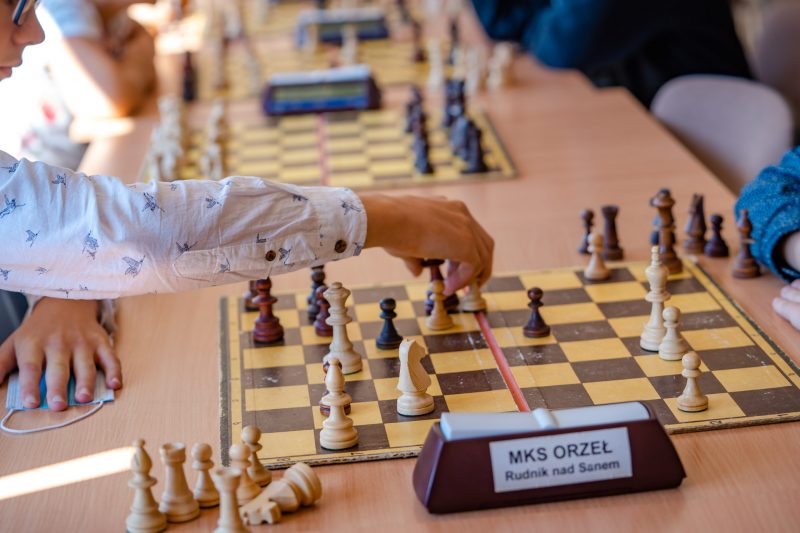 Szachista po lewej stronie stolika wykonuje ruch na szachownicy. Na pierwszym planie zegar z napisem MKS Orzeł Rudnik nad Sanem