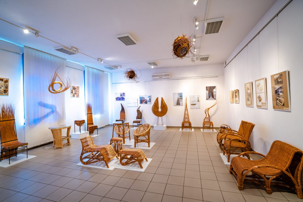 Oryginalne meble i formy z wikliny ustawiony na postumencie w sali wystawowej. Na ścianach kolorowe zdjęcia