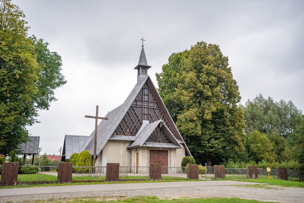 Mały kościółek ze spadzistym dachem i portalem ze szkła i drewna. Przy kościele krzyż