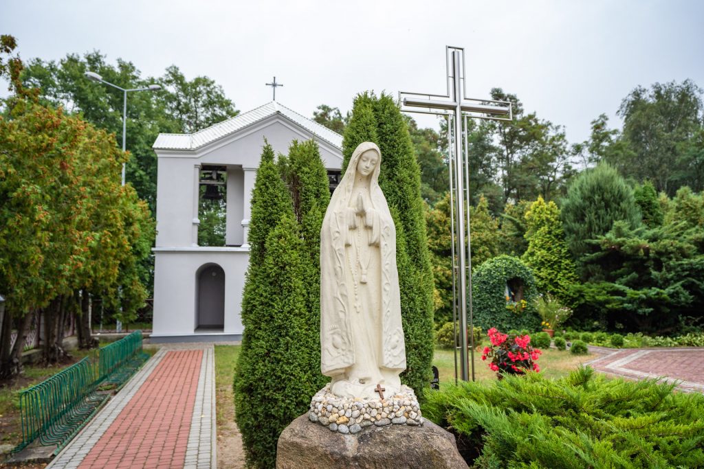 Fifura Matki Boskiej z jasnego kamienia, w tle cyprysy, krzyż i biała dzwonnica