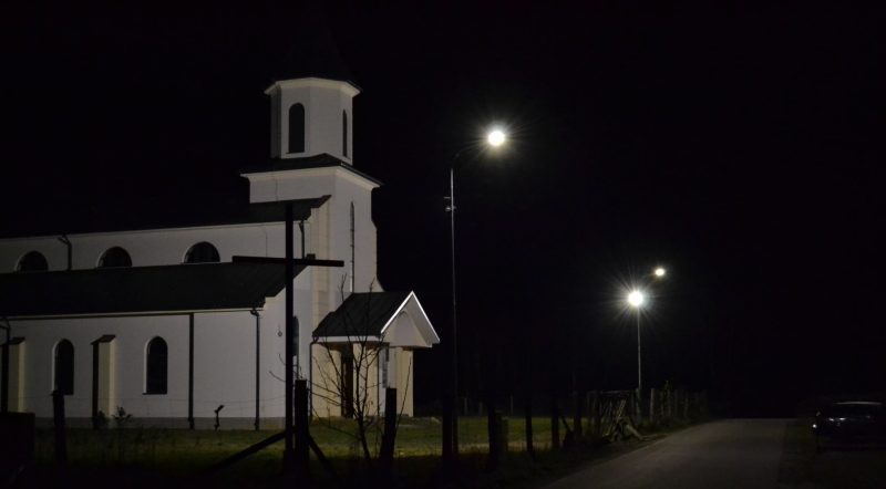 Iluminowany kościół z wieżą i jasną elewacją. Przed kościołem dwie zapalone latarnie